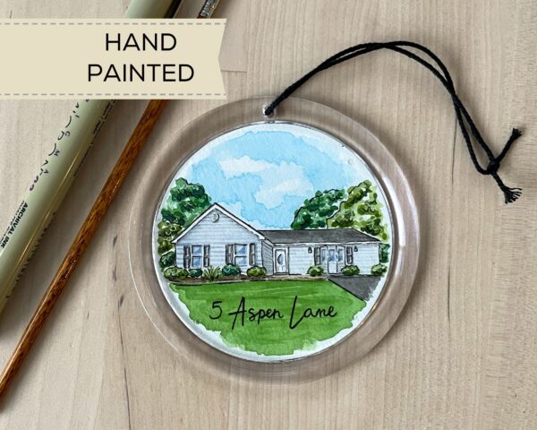 A clear round ornament with a hand painted watercolor painting of a blue house inside of it and the address written along the bottom.