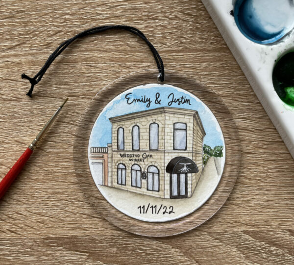 A clear round ornament with a hand painted watercolor painting of a wedding venue inside of it.