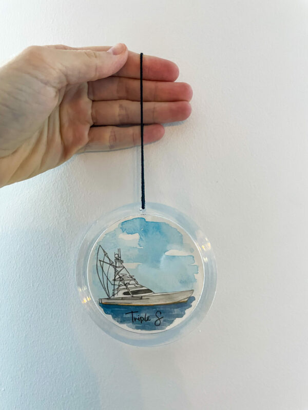 A hand holding a clear round ornament with a hand painted watercolor painting of a boat inside of it.