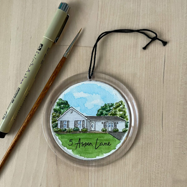 A clear round ornament with a hand painted watercolor painting of a blue house inside of it.