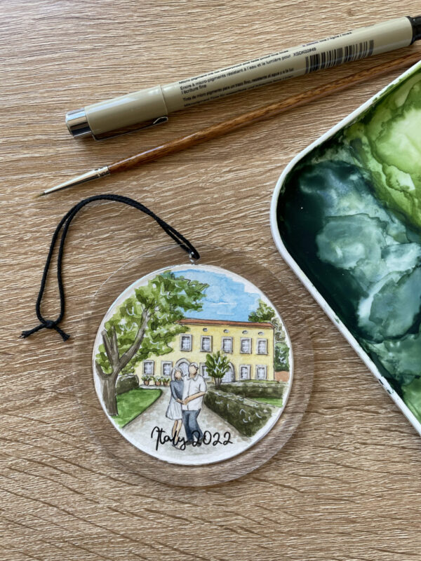 A clear round ornament with a hand painted watercolor painting of a couple standing in front of an Italian villa inside of it.