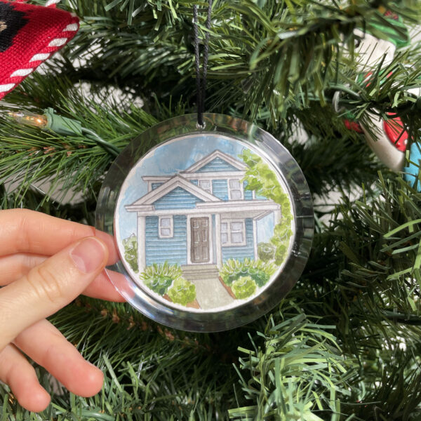 A clear round ornament with a hand painted watercolor painting of a blue house inside of it hanging from a Christmas Tree.