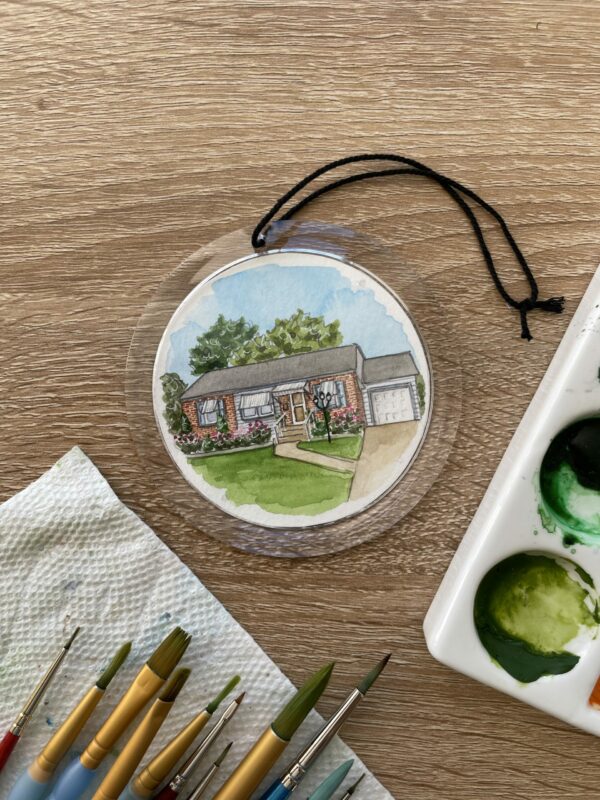 A clear round ornament with a watercolor painting of a brick house inside of it.