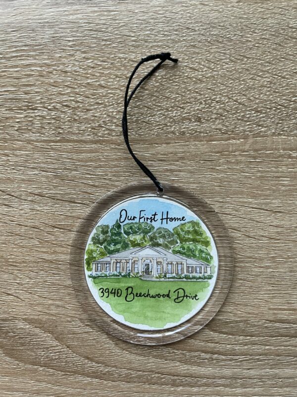 A clear round ornament with a watercolor painting of a house inside of it. The words "our first home" are written along the top of the painting with the address written below.