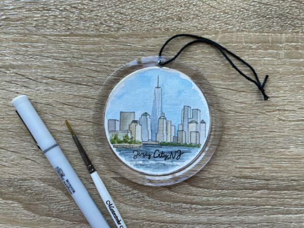 A clear round ornament with a watercolor painting of the skyline of downtown New York City inside of it.