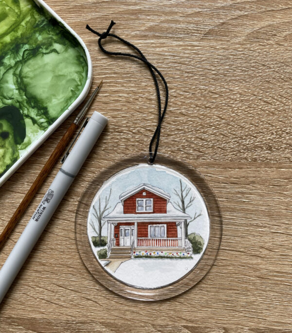 A clear round ornament with a hand painted watercolor painting of a red house inside of it. The house is covered in snow with Christmas lights.